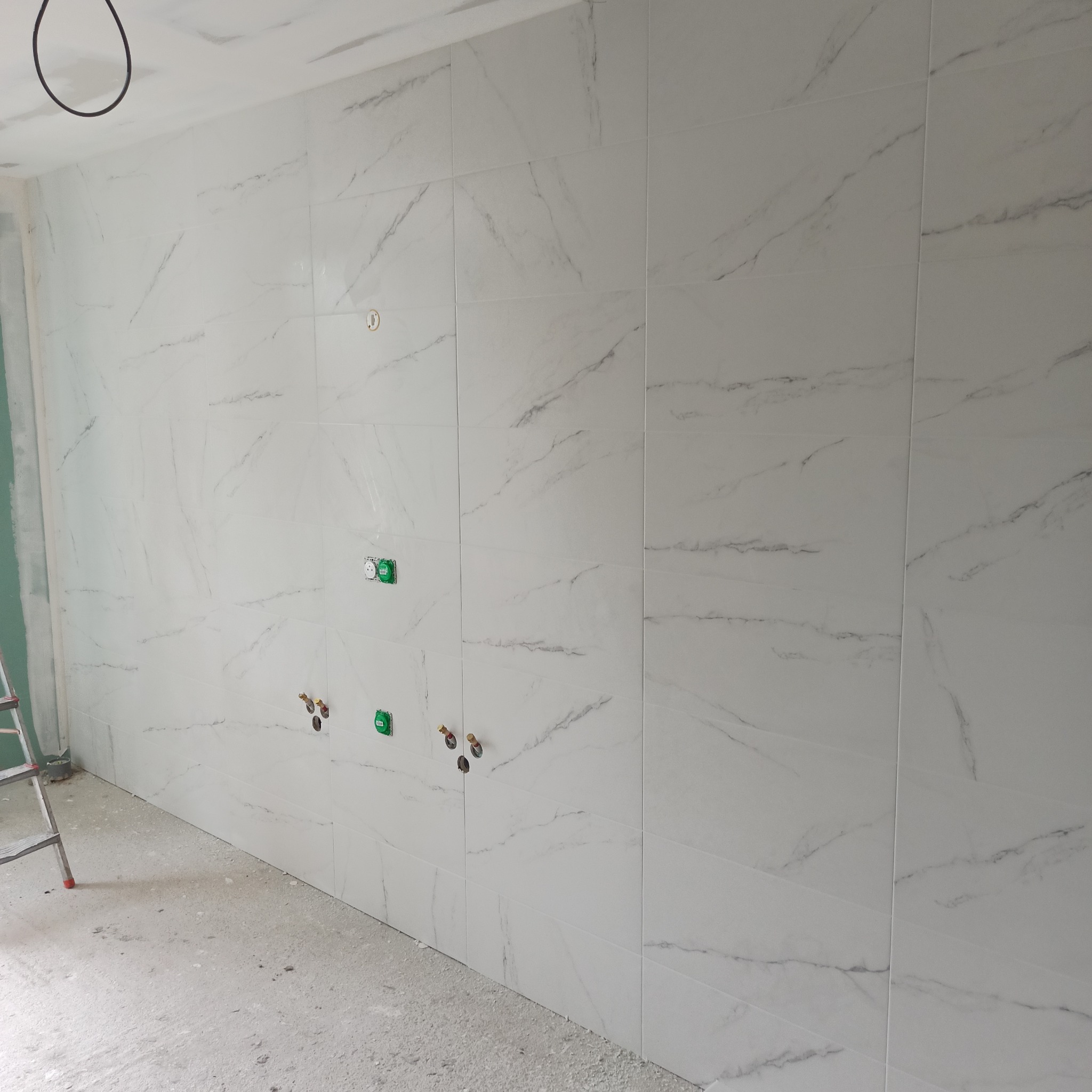 Wykończona ściana w łazience z białych płytek imitujących marmur, widoczne gniazdka i wyprowadzenia hydrauliczne. Na podłodze resztki zaprawy, w rogu drabina.