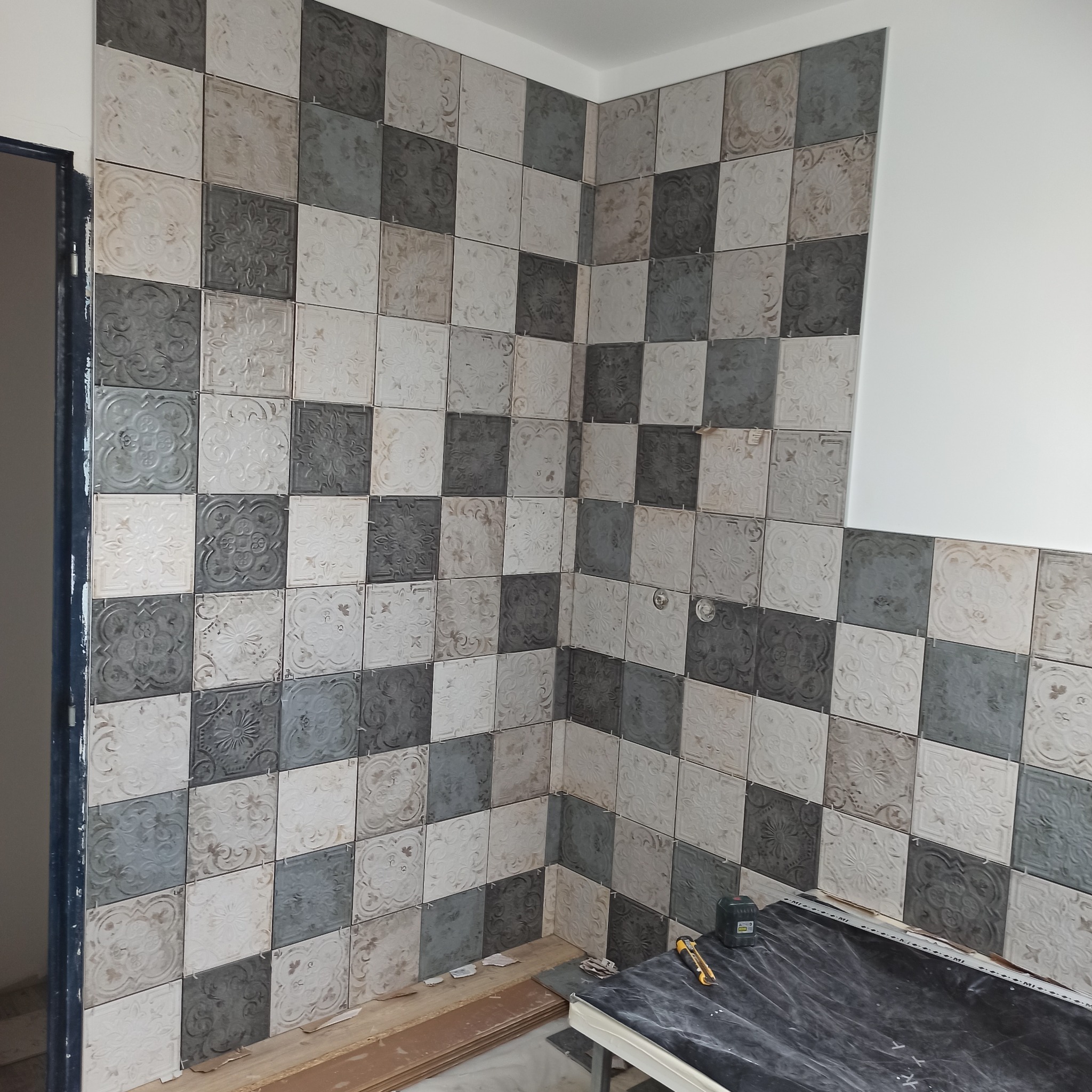 Ściana wyłożona dekoracyjnymi, kwadratowymi płytkami w odcieniach szarości i beżu. Widoczne narzędzia glazurnicze i fragment niezabudowanej ściany.