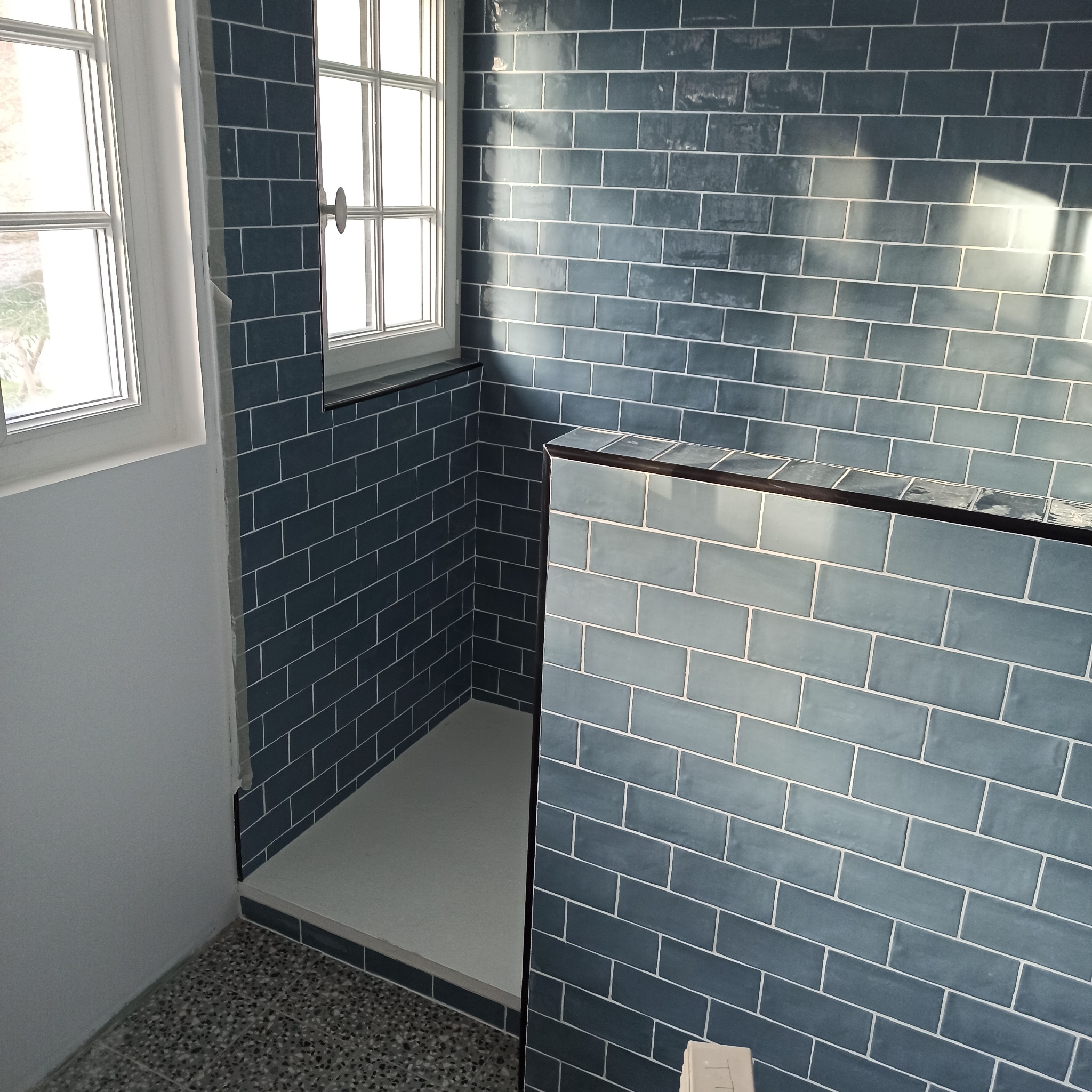 Nisko: Kabina prysznicowa z niebieskimi, błyszczącymi płytkami ułożonymi w cegiełkę, z widocznym oknem i brodzikiem. Nowoczesny design.