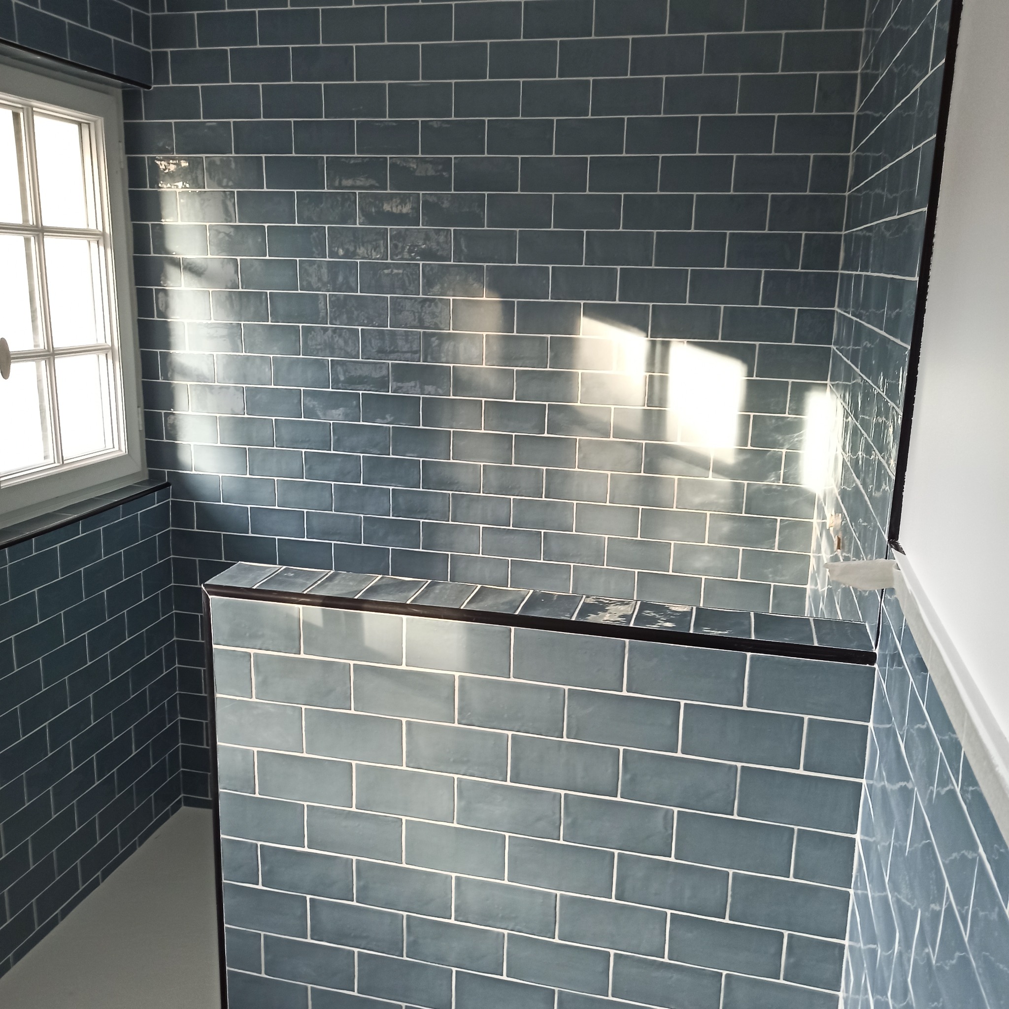 Błękitne, prostokątne płytki w łazience z białymi fugami, ułożone w cegiełkę. Widoczne okno z białymi ramami i fragment ściany z listwą przypodłogową.