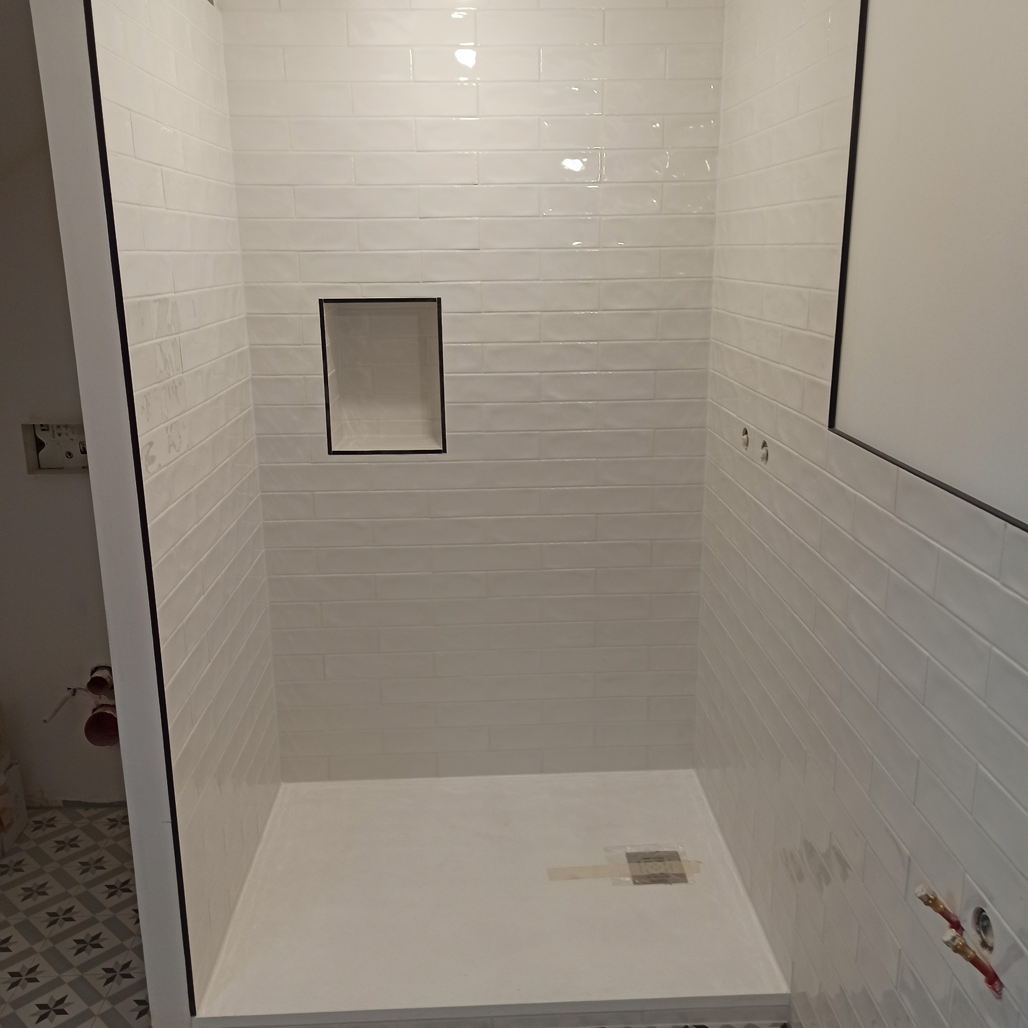 Biała kabina prysznicowa wyłożona błyszczącymi, prostokątnymi płytkami z czarną ramką wnęki. Widoczne przyłącza wodne i fragment podłogi z wzorzystymi płytkami.