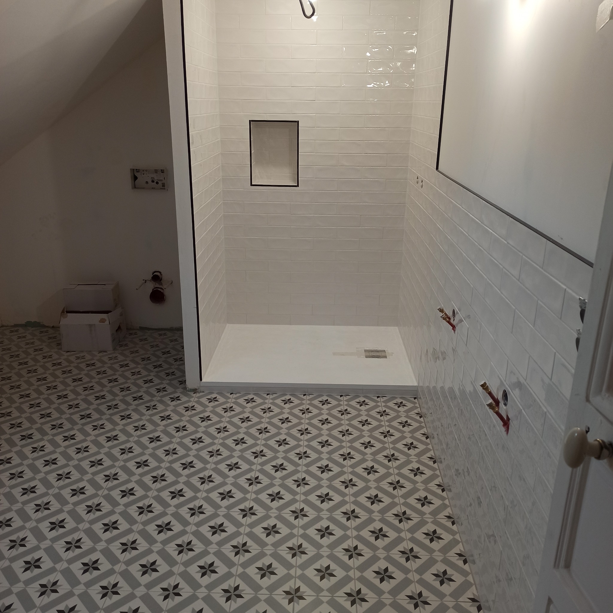 Biała łazienka w trakcie wykończenia: kabina prysznicowa z wnęką, wzorzysta podłoga, widoczne instalacje. Ujęcie z perspektywy wejścia, surowe ściany w tle.