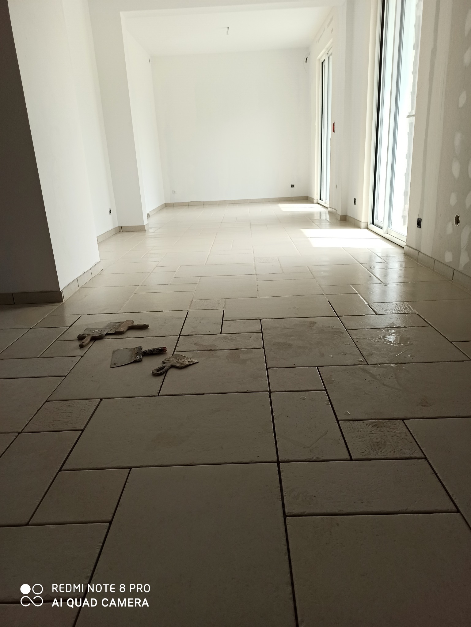 Jasne, prostokątne płytki podłogowe ułożone w nowym, pustym pomieszczeniu. Widoczne narzędzia glazurnicze. Duże okno w tle. Świeżo położona podłoga.