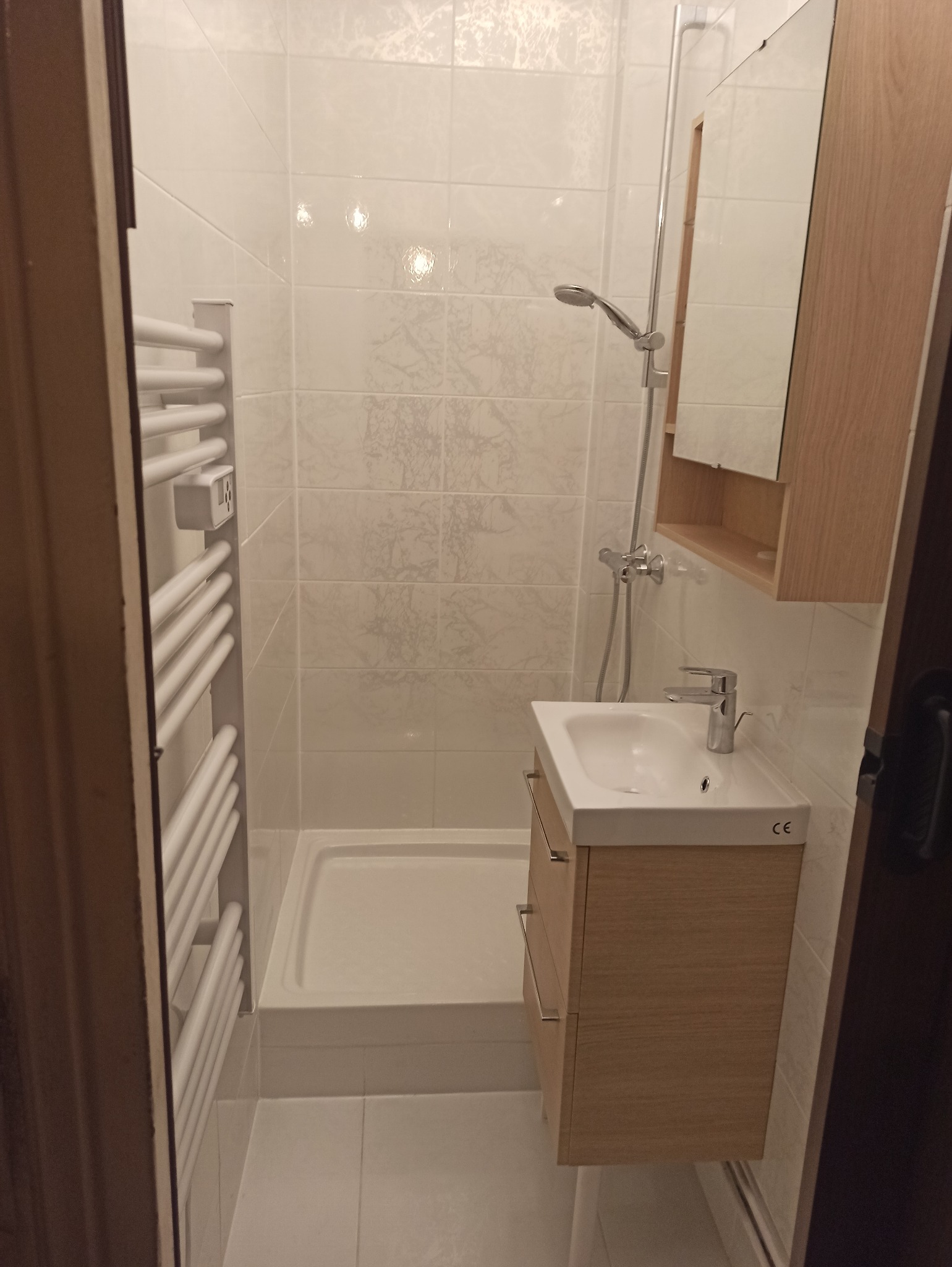 Jasna łazienka po remoncie: białe płytki z delikatnym wzorem, kabina prysznicowa, umywalka z szafką z jasnego drewna i grzejnik drabinkowy. Nowoczesny design.