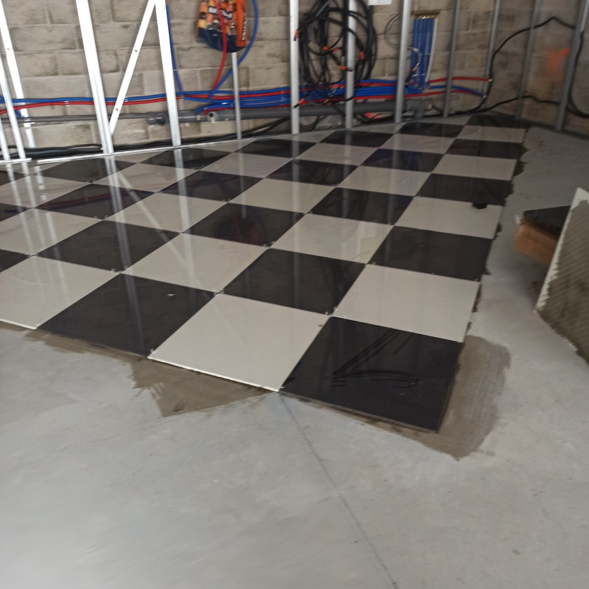 Ułożone płytki podłogowe w szachownicę, widoczne instalacje w tle. Etap prac wykończeniowych w pomieszczeniu z surowymi ścianami i podłogą.