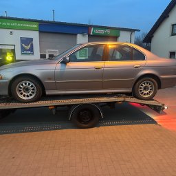 Tomasz Simlat - Szary sedan BMW E39 na lawecie, na tle warsztatu samochodowego z logo Castrol i F Auto Artur Fuhrmann. Widoczne światła pozycyjne lawety.