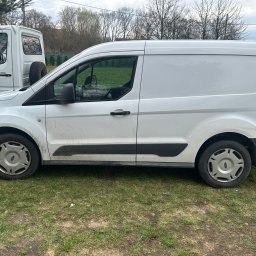 Tomasz Simlat - Biały, lekko zabrudzony furgon Ford Transit Connect na trawie, widziany z boku. W tle inny biały pojazd transportowy i drzewa bez liści.