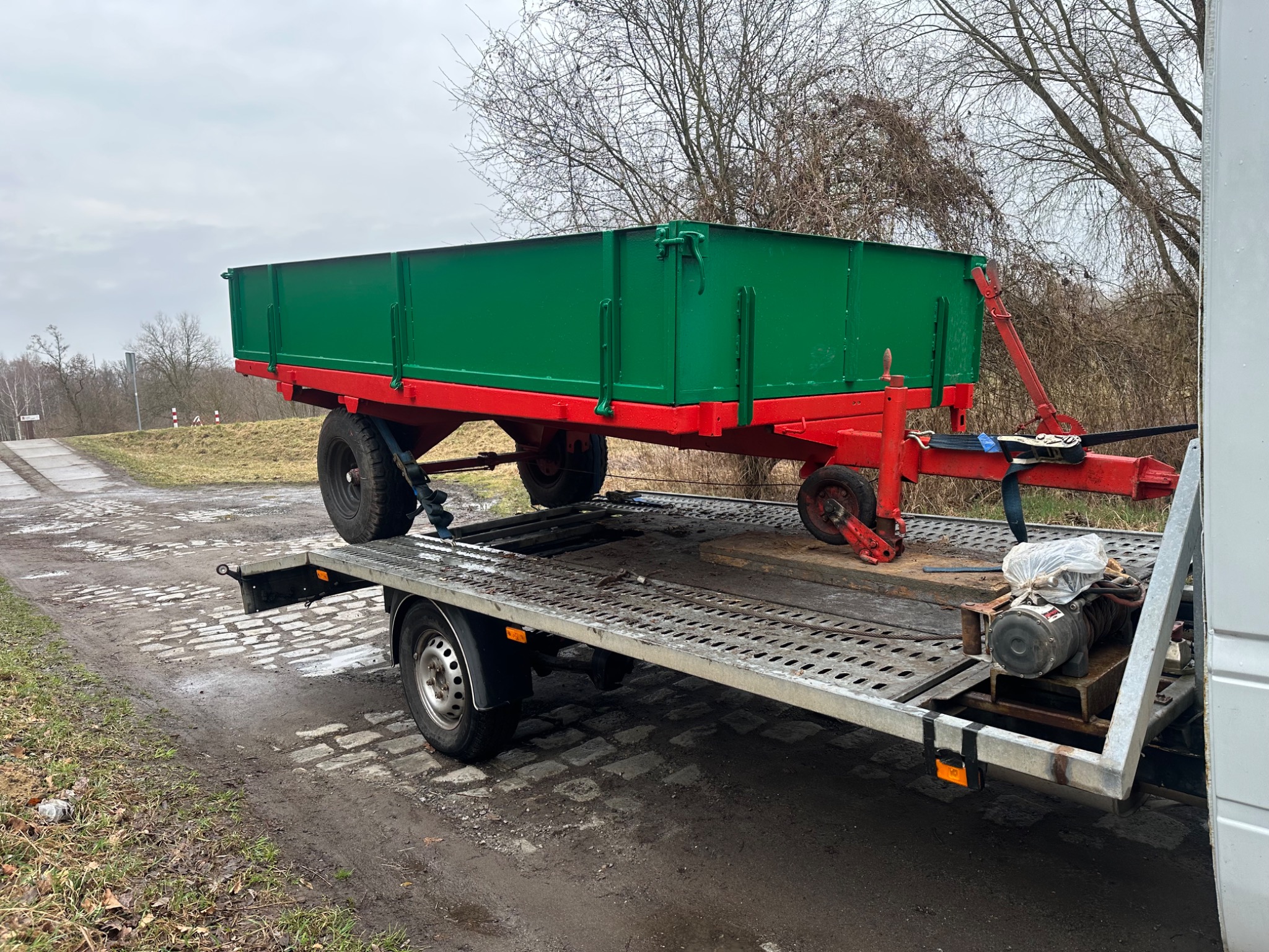 Zielono-czerwona przyczepa rolnicza na ażurowej lawecie transportowej, widok z boku na tle drzew bez liści i szarego nieba. Detal: wyciągarka na lawecie.