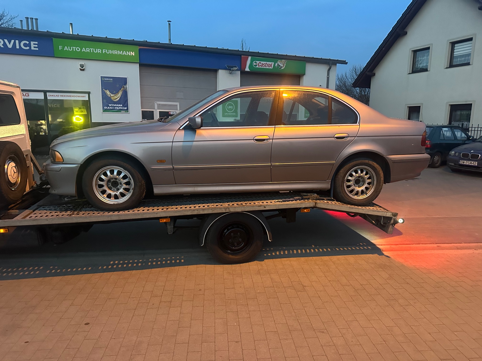 Szary sedan BMW E39 na lawecie, na tle warsztatu samochodowego z logo Castrol i F Auto Artur Fuhrmann. Widoczne światła pozycyjne lawety.