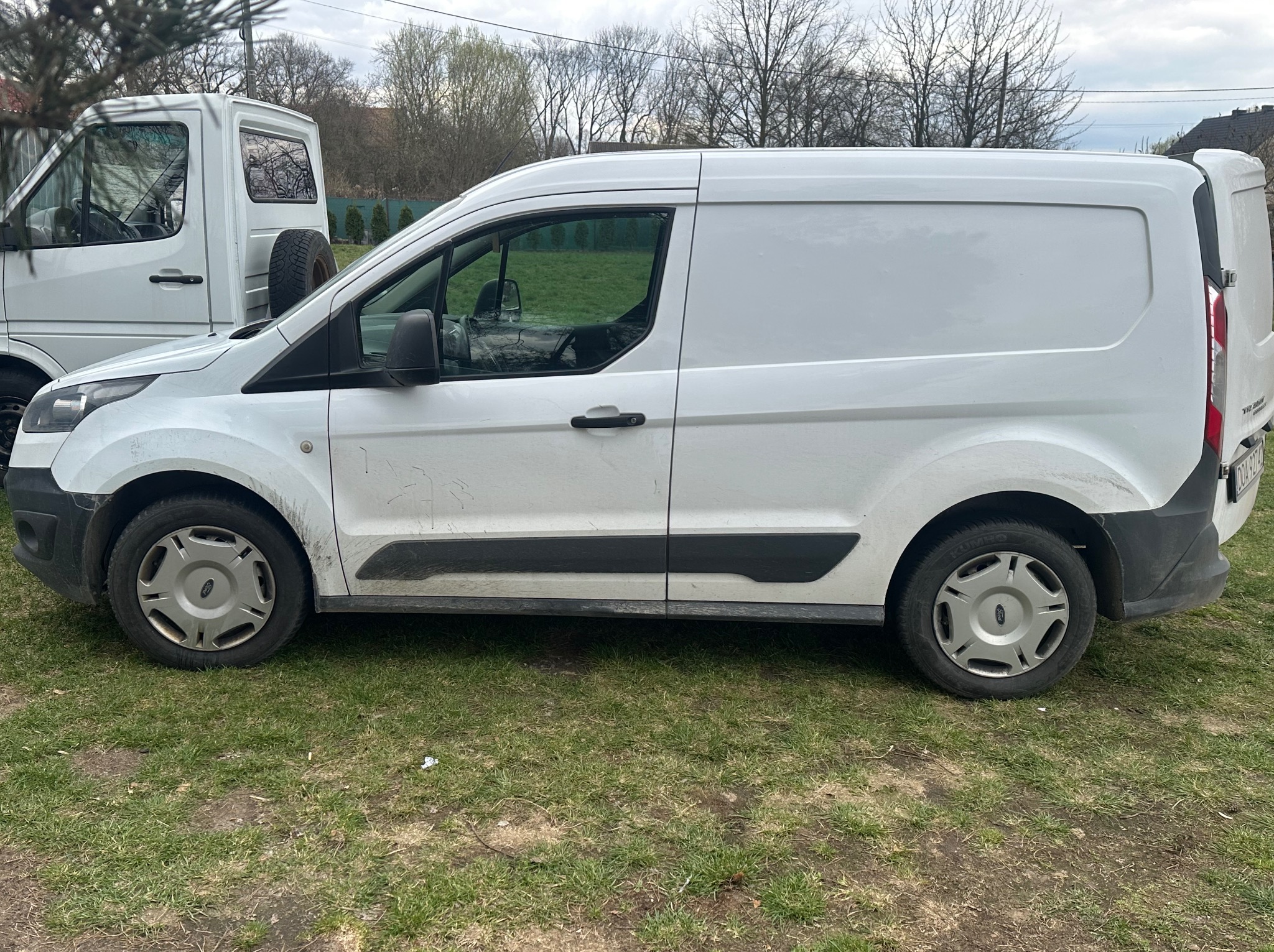 Biały, lekko zabrudzony furgon Ford Transit Connect na trawie, widziany z boku. W tle inny biały pojazd transportowy i drzewa bez liści.