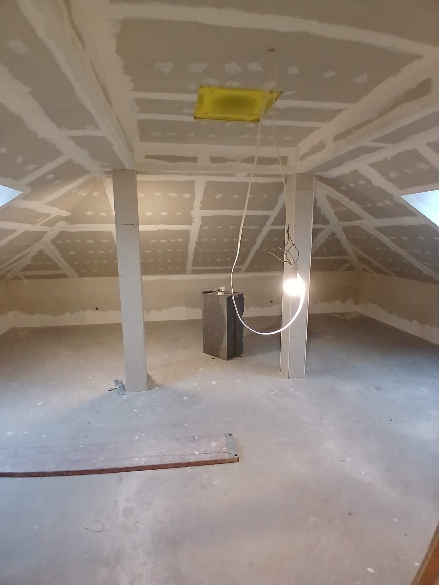Poddasze w trakcie prac wykończeniowych: płyty karton-gips na skosach i suficie, widoczne łączenia do szpachlowania, surowa podłoga, oświetlenie budowlane.