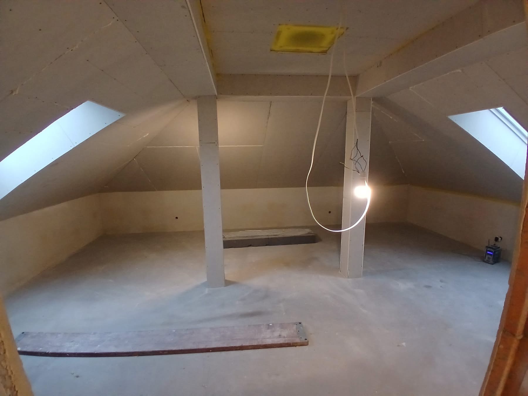 Poddasze z oknami dachowymi i płytami kartonowo-gipsowymi na ścianach i suficie, widoczne słupy konstrukcyjne i fragment podłogi. Prace wykończeniowe w trakcie.