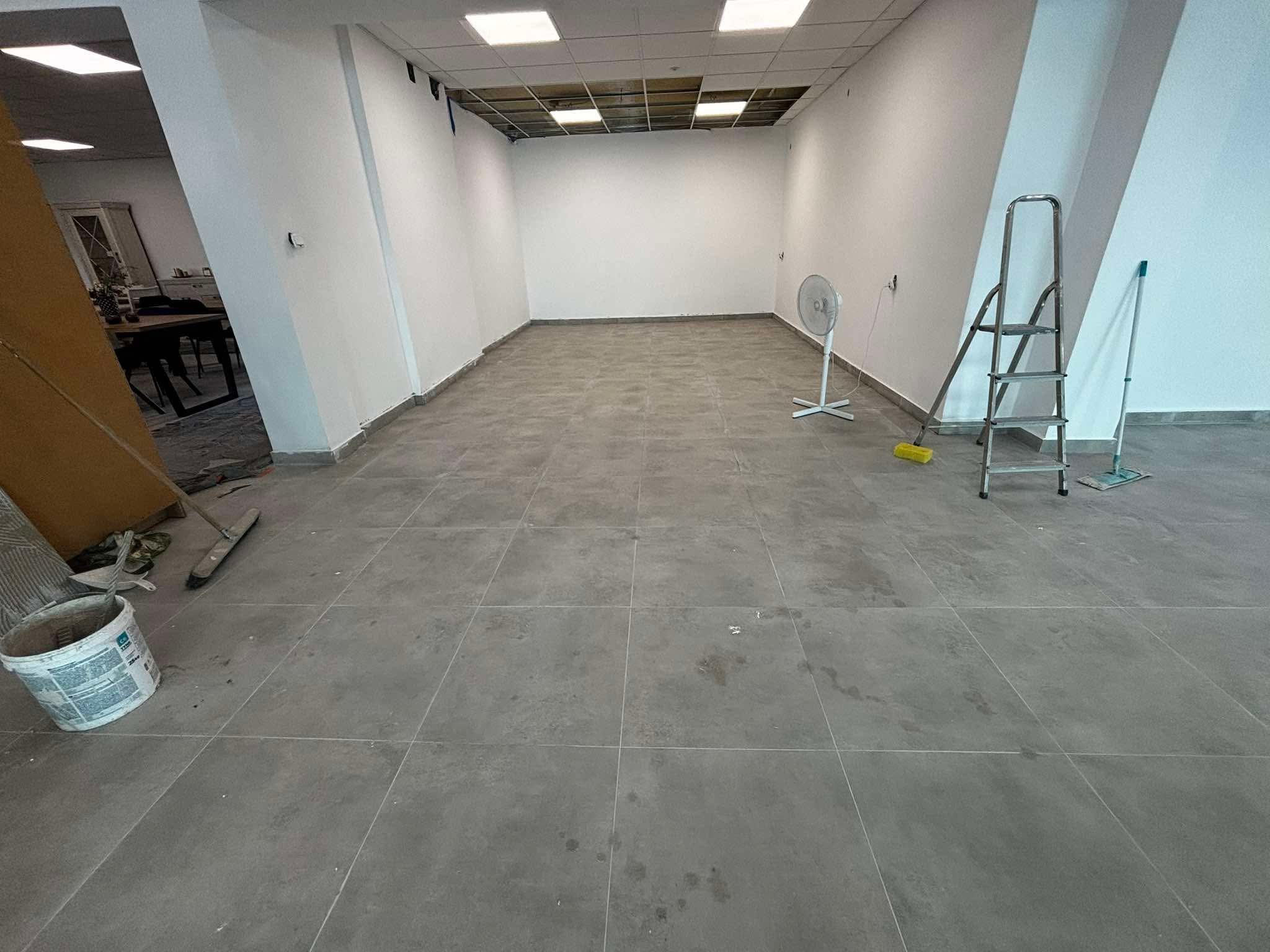 Wnętrze biura po malowaniu ścian na biało, widoczna drabina, wentylator i wiadro z farbą. Szare płytki na podłodze. Sufit z odsłoniętą instalacją.