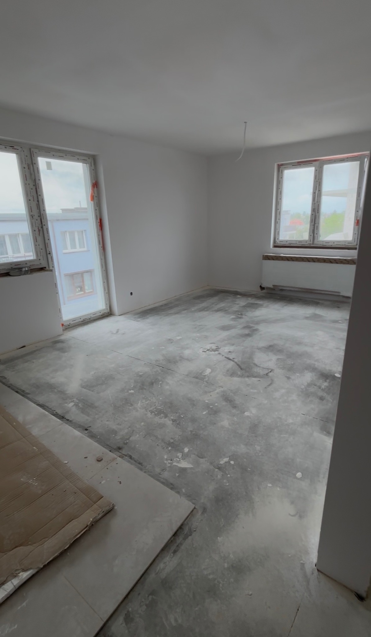 Wnętrze pokoju w trakcie remontu: białe ściany, dwa okna z widokiem na budynek, surowa podłoga z widocznymi śladami prac, karton na podłodze.