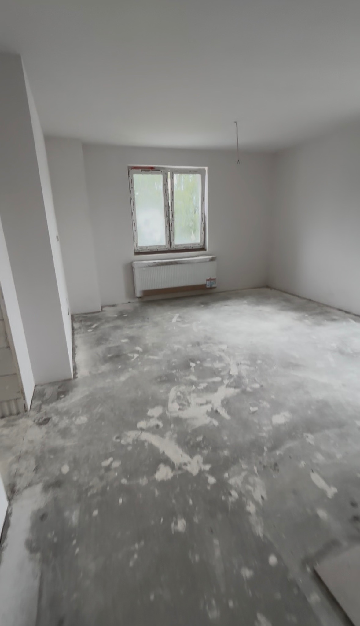 Pokój w trakcie remontu: surowe ściany, okno z widokiem na zieleń, grzejnik. Podłoga z pozostałościami po pracach budowlanych. Widoczne niedoskonałości i surowość wnętrza.