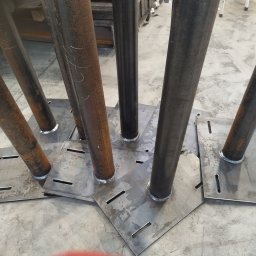 Magstal - Metalowe słupy z przyspawanymi podstawami, widoczne spawy i otwory w podstawach. Część słupów rdzawa, część pomalowana na czarno. Szary beton na podłożu.