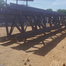 Magstal - Stalowa konstrukcja kratowa hali w trakcie montażu, widoczna na placu budowy z zielonym podnośnikiem teleskopowym w tle, oświetlona jasnym światłem słonecznym.