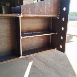 Magstal - Fragment konstrukcji stalowej z widocznymi spawami i otworami, ustawionej na betonowej posadzce w hali. Widoczne elementy zardzewiałego metalu i ślady po spawaniu.