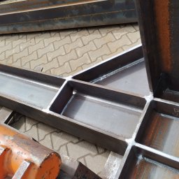 Magstal - Spojrzenie z góry na spawane, stalowe profile konstrukcyjne, leżące na brukowanej powierzchni. Widoczne ślady rdzy i pomarańczowy element hydrauliczny.