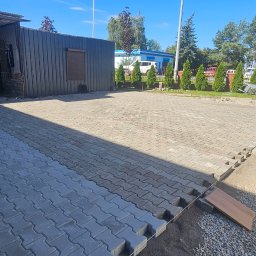 Trans Europe Artur Wróbel - Fragment układanej kostki brukowej na podjeździe, z widocznymi pracownikami i narzędziami. Część powierzchni już wyłożona, reszta w trakcie prac. Słoneczny dzień.