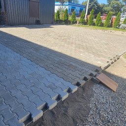 Trans Europe Artur Wróbel - Częściowo ułożona, szara kostka brukowa na podjeździe. Widoczny fragment podsypki, narzędzia pracy i butelka napoju. W tle budynek i zieleń.