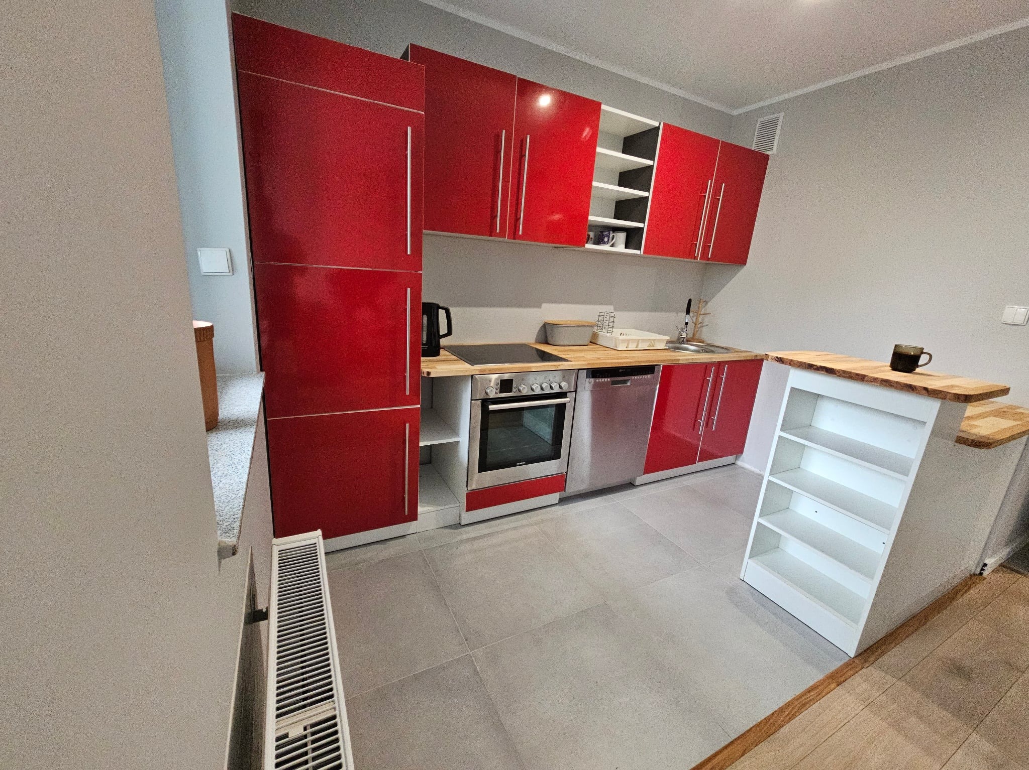 Nowoczesna kuchnia z czerwonymi szafkami, drewnianym blatem i szarymi płytkami podłogowymi. Widoczny piekarnik, zlew i półka barowa. Minimalistyczny design.