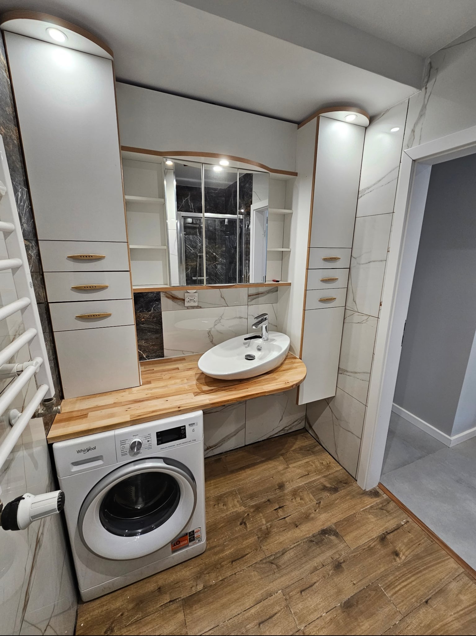 Nowoczesna łazienka z drewnianym blatem, umywalką nablatową i pralką. Białe szafki z drewnianymi akcentami, marmurowe płytki i drewnopodobna podłoga tworzą spójną całość.