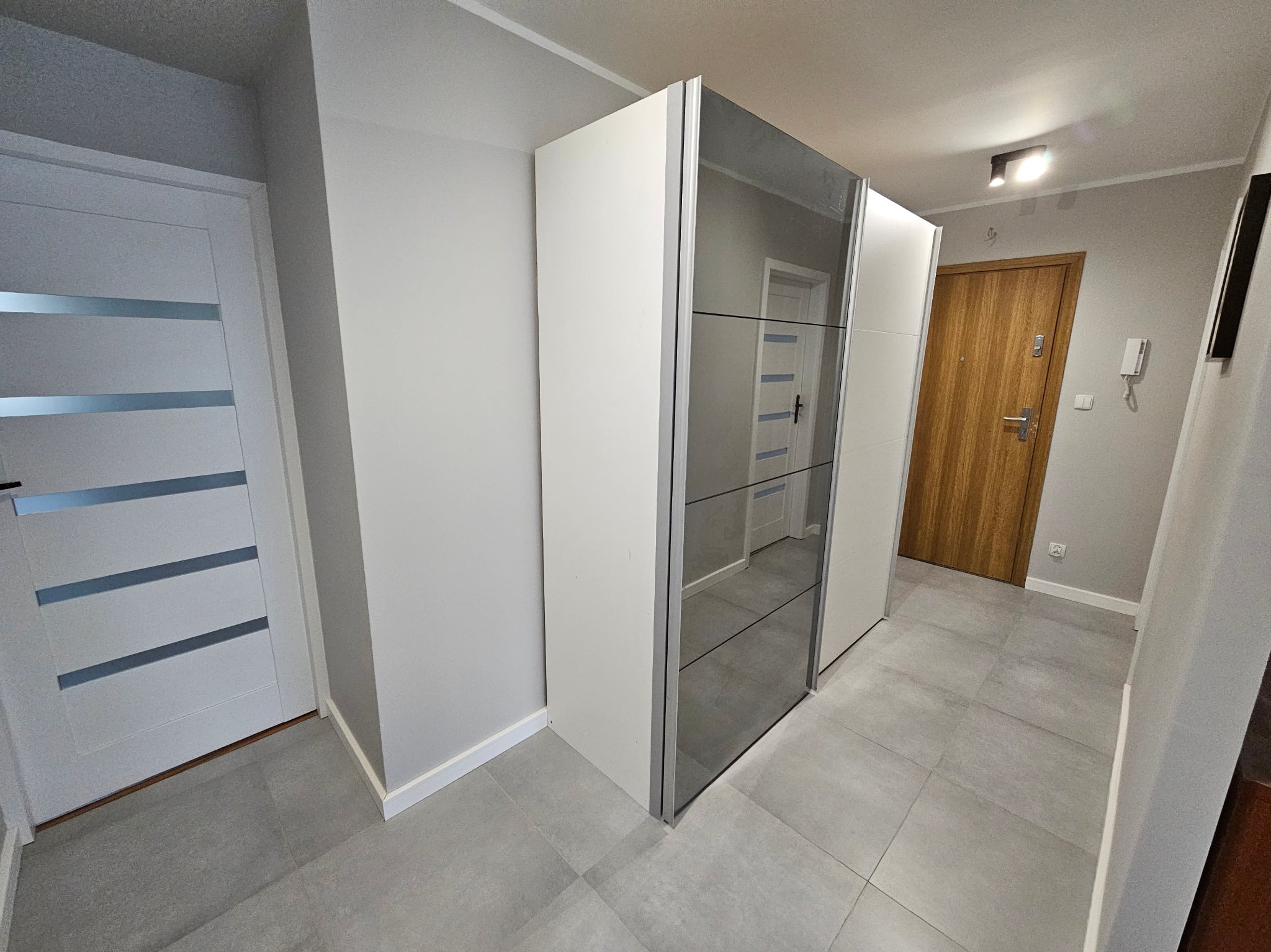 Nowoczesny przedpokój z szarą podłogą, białą szafą z lustrzanymi drzwiami i białymi drzwiami z poziomymi przeszkleniami. Widoczne oświetlenie sufitowe i drewniane drzwi.