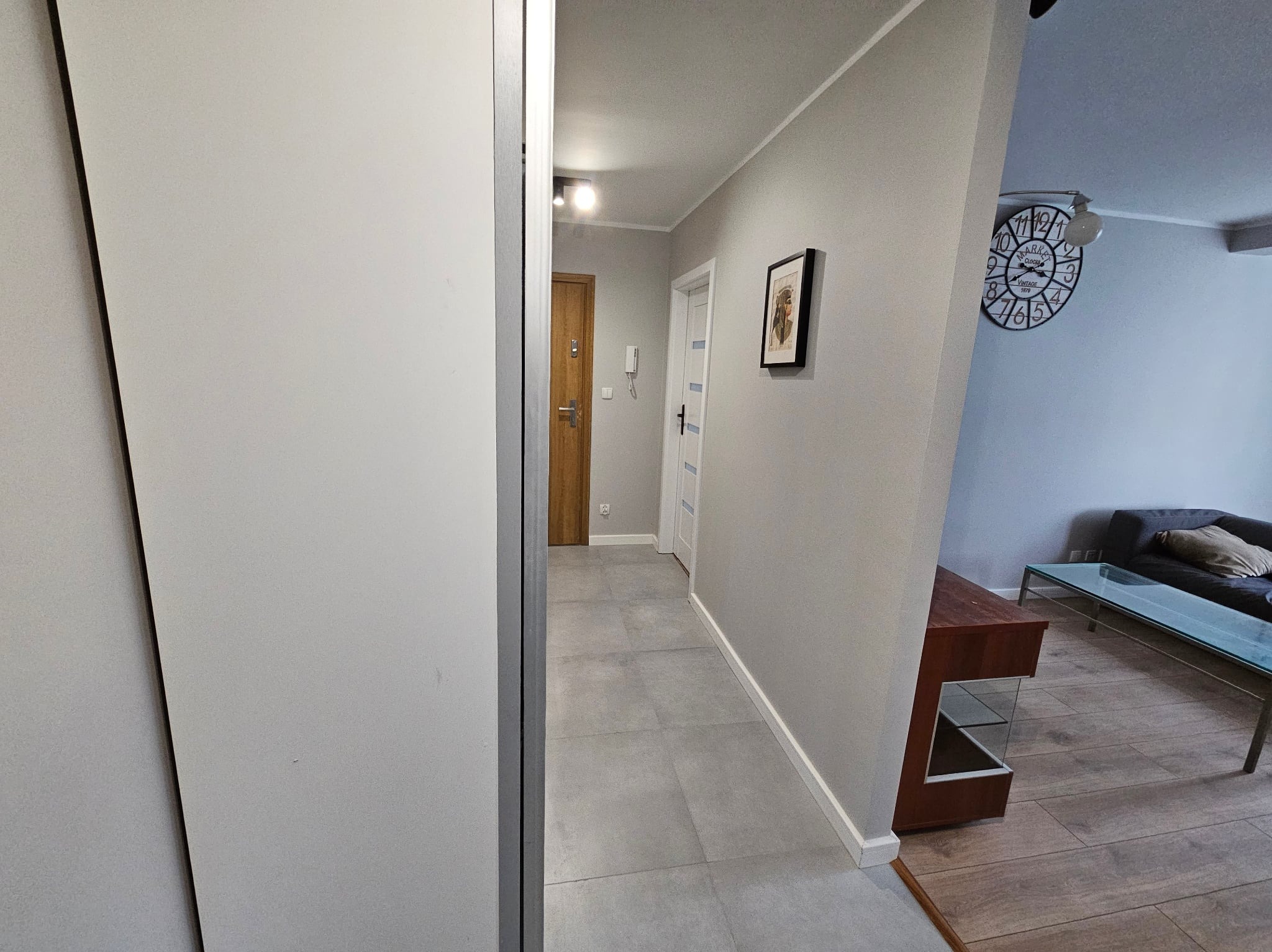 Jasne wnętrze mieszkania z szarą podłogą i ścianami. Widoczny fragment szafy, drzwi wejściowe i salon z zegarem na ścianie. Minimalistyczny design.