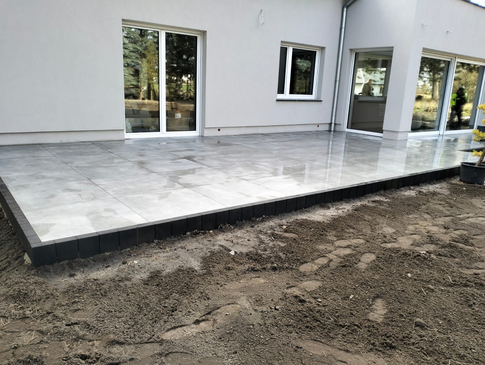 Nowoczesny taras z szarych płyt, obrzeża z ciemnej kostki. Widoczne przesuwne drzwi tarasowe i fragment ogrodu. Świeżo ułożona kostka, ziemia wokół.