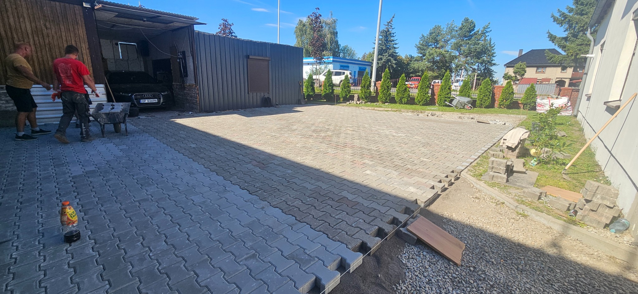 Fragment układanej kostki brukowej na podjeździe, z widocznymi pracownikami i narzędziami. Część powierzchni już wyłożona, reszta w trakcie prac. Słoneczny dzień.