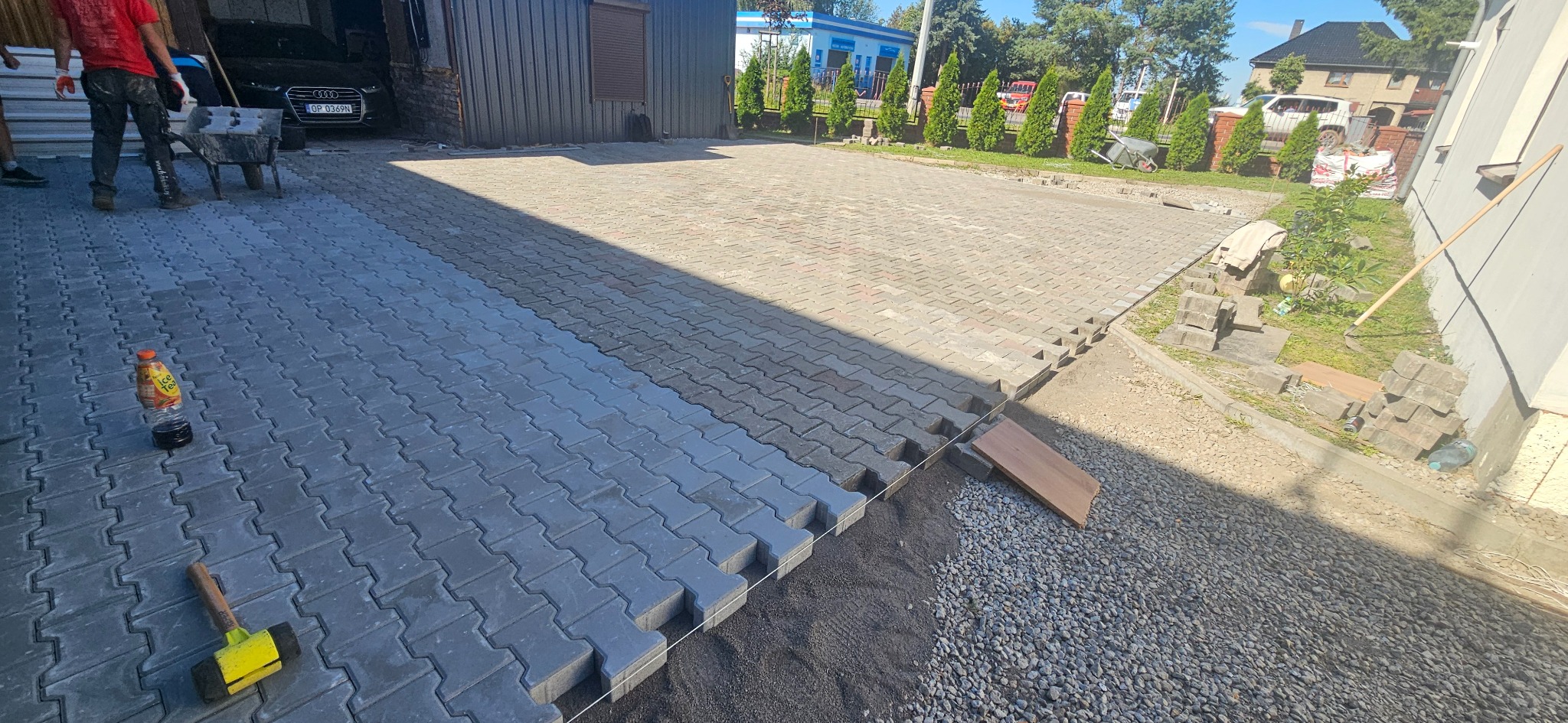 Częściowo ułożona, szara kostka brukowa na podjeździe. Widoczny fragment podsypki, narzędzia pracy i butelka napoju. W tle budynek i zieleń.