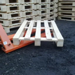 Świeża, jasna paleta drewniana na pomarańczowym wózku widłowym, w tle stosy palet na czarnym podłożu.