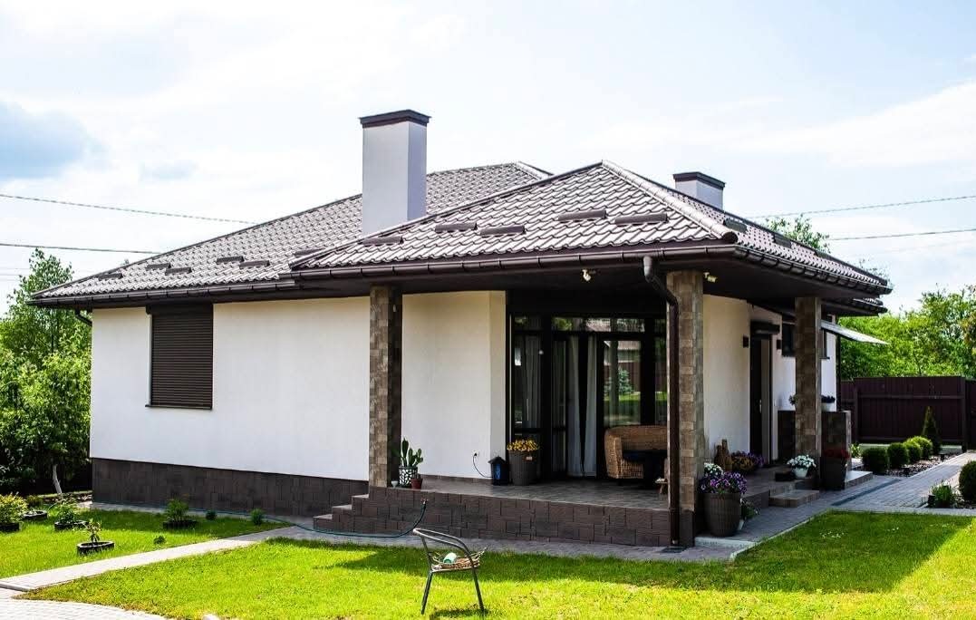 Parterowy dom z brązowym dachem i kolumnami z imitacji kamienia. Widoczny taras z meblami ogrodowymi i zadbany trawnik. Dom w stylu nowoczesnym.
