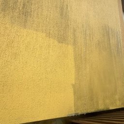 HydroEfekt - Żółta elewacja budynku z wyraźnym kontrastem czystej i zabrudzonej powierzchni, ukazując efekt mycia. Widoczny fragment kamiennej podmurówki i okapu.