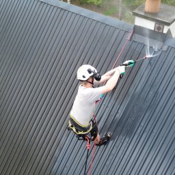 HydroEfekt - Pracownik w kasku, na linie, myje ciemny, blaszany dach z użyciem myjki ciśnieniowej. Widok z góry, w tle komin i zieleń.