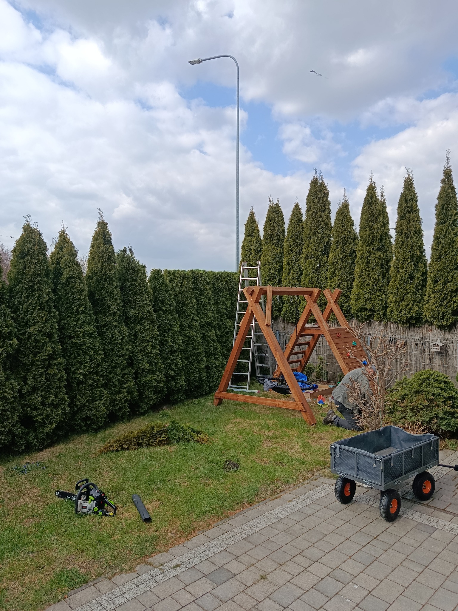 Montaż drewnianego placu zabaw w ogrodzie w Łomży. W tle rząd tuj, na trawie leży piła łańcuchowa, obok stoi wózek transportowy. Prace ogrodowe w toku.