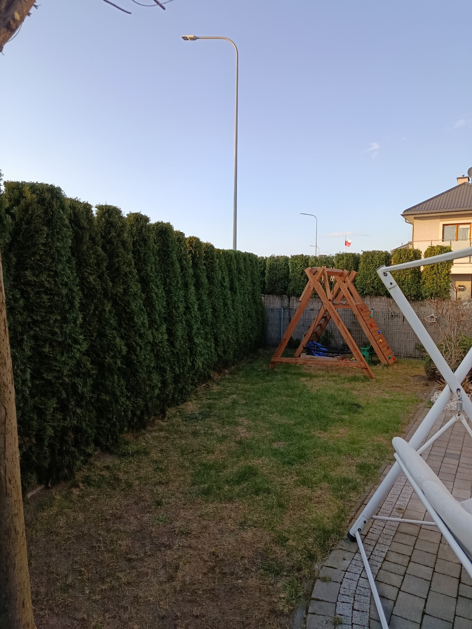 Zadbany ogród z równo przyciętym żywopłotem z tui, drewnianym placem zabaw i huśtawką. Widoczne oświetlenie i fragment domu w tle.