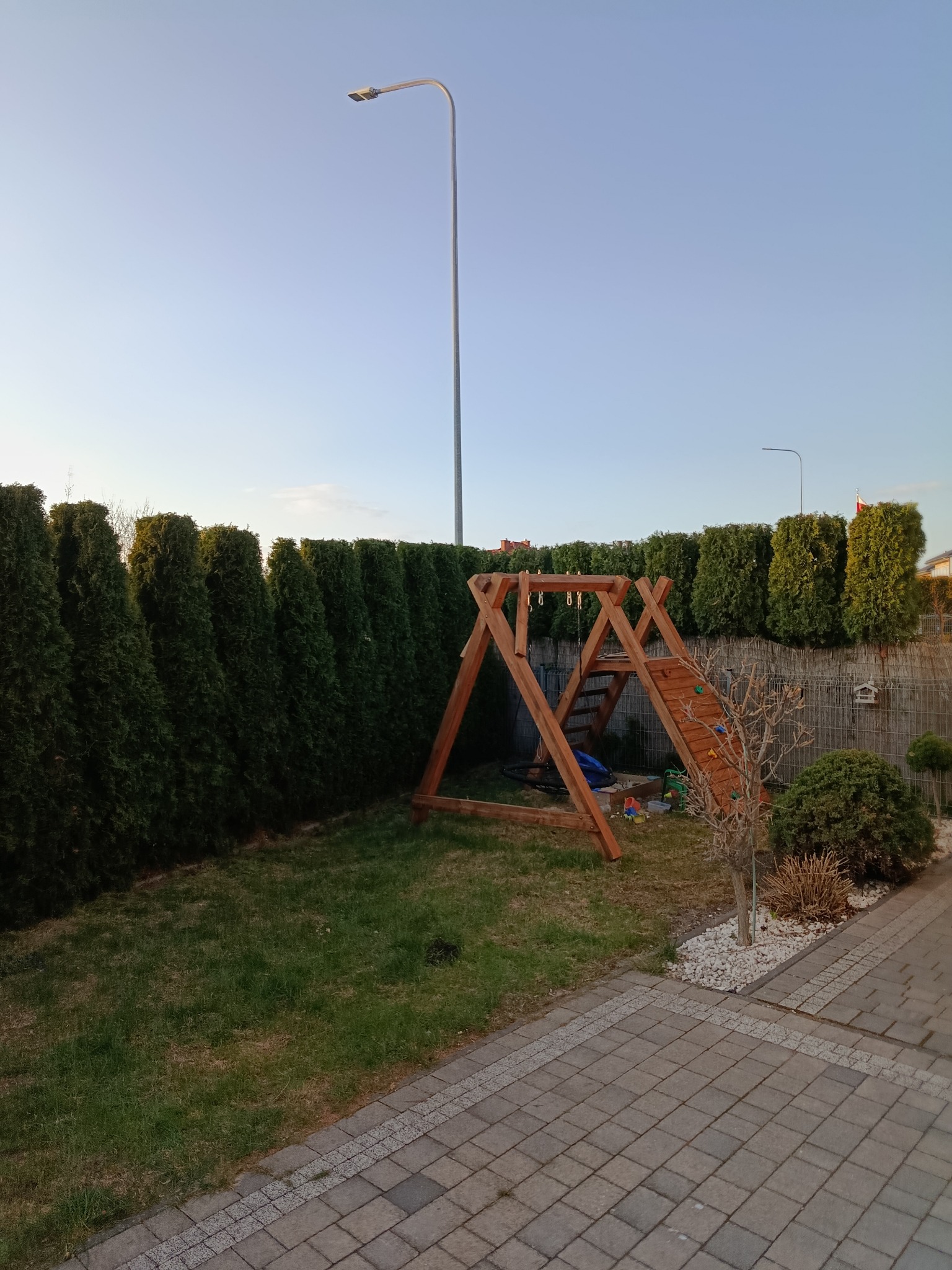 Zadbany ogród z równo przyciętymi tujami wzdłuż ogrodzenia. Na trawniku drewniany plac zabaw dla dzieci. Widoczne oświetlenie ogrodowe i kostka brukowa.