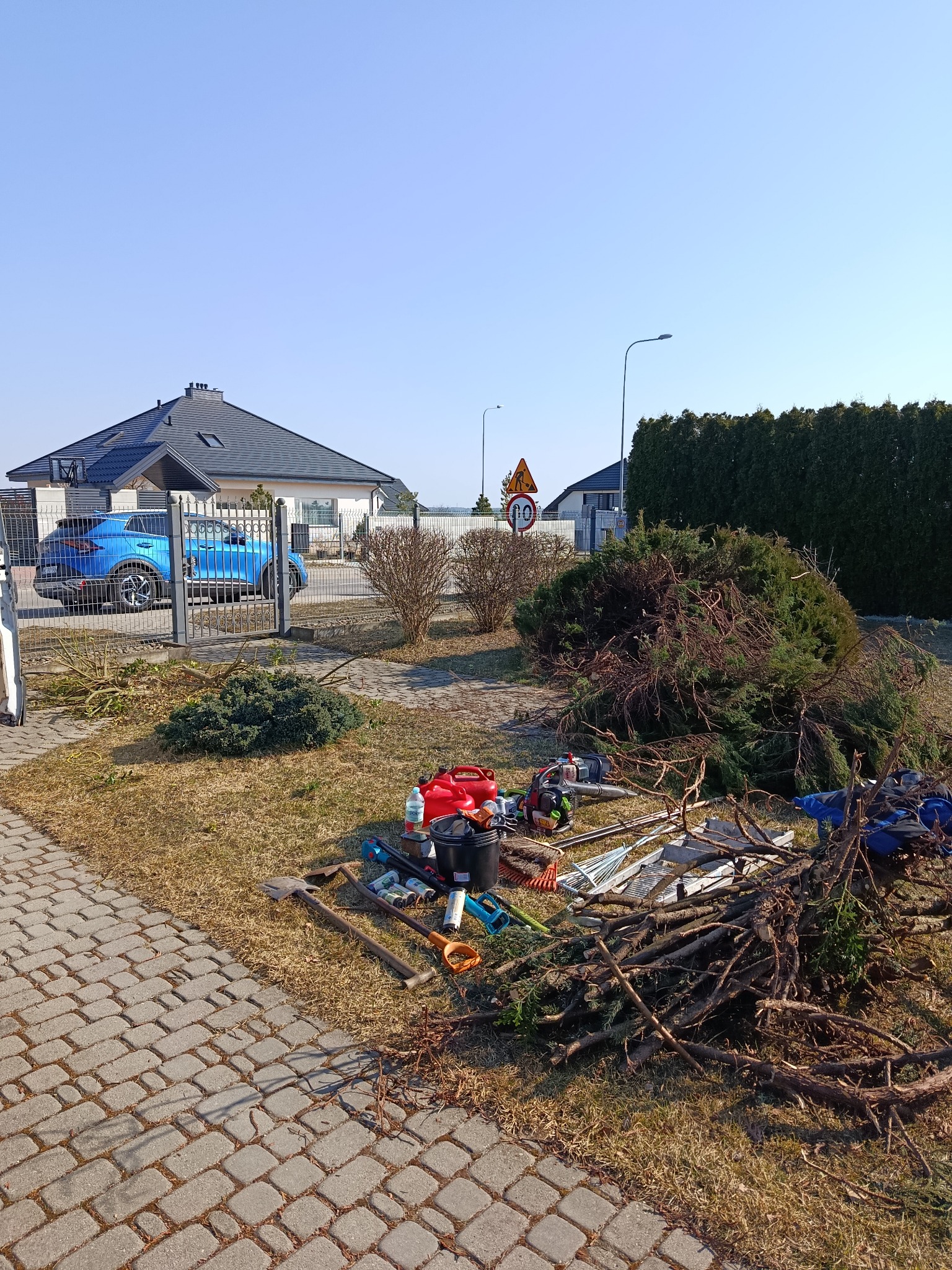 Stos gałęzi po przycince krzewów i narzędzia ogrodnicze na trawniku przed domem z niebieskim autem. Widok na uporządkowany teren po pracach ogrodowych.