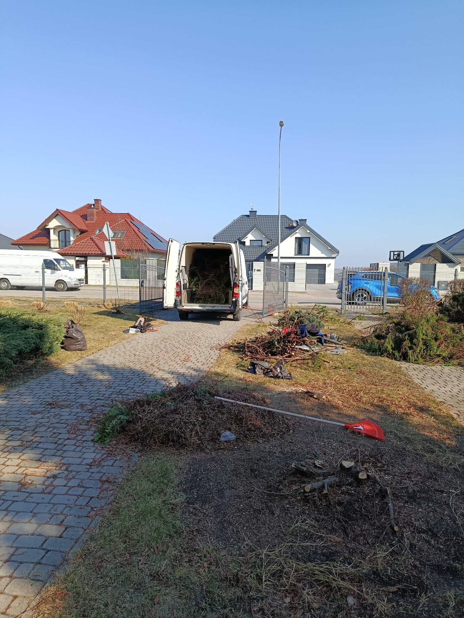 Otwarty van wypełniony gałęziami na tle domów. Na podjeździe sterta gałęzi, grabie i torba. Widoczne ślady po wycince drzew. Słoneczny dzień.