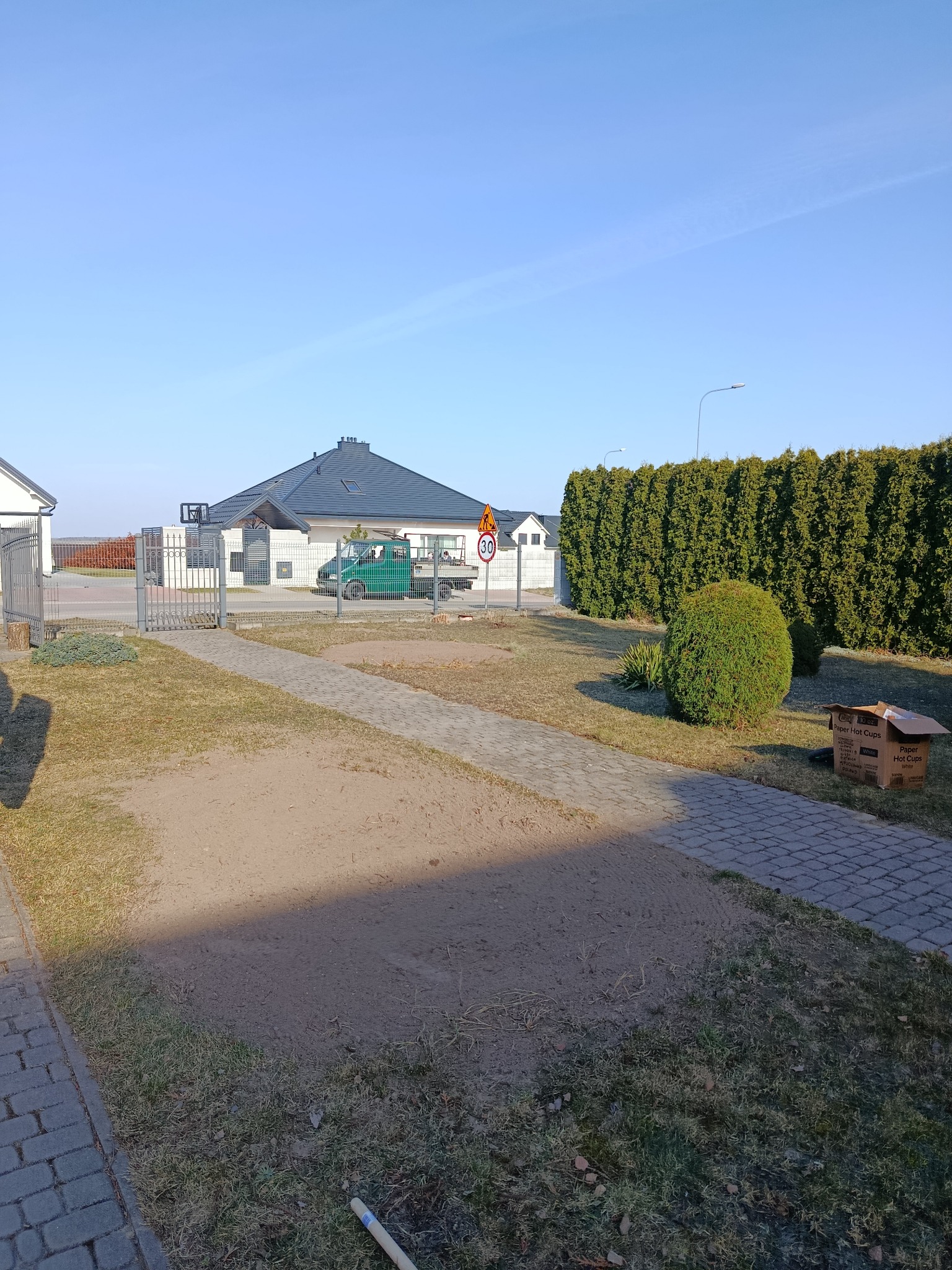 Przygotowany teren pod trawnik z widocznym samochodem dostawczym, krzewem i żywopłotem w tle. Świeżo wyrównana ziemia czeka na zasianie lub ułożenie trawy.