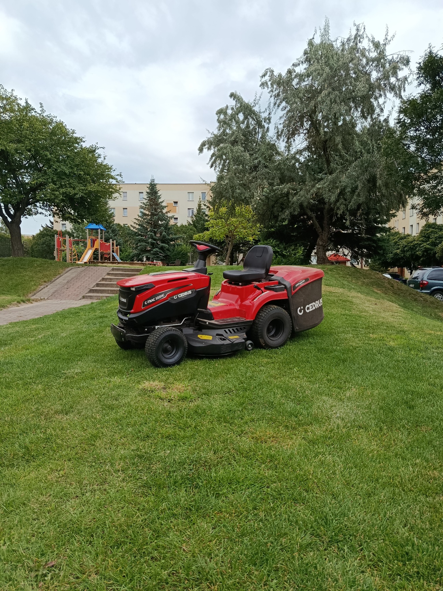 Czerwona kosiarka traktorek Cedrus na trawniku, w tle blok, drzewa i plac zabaw. Sprzęt po koszeniu, trawa równo skoszona. Widok na osiedle.