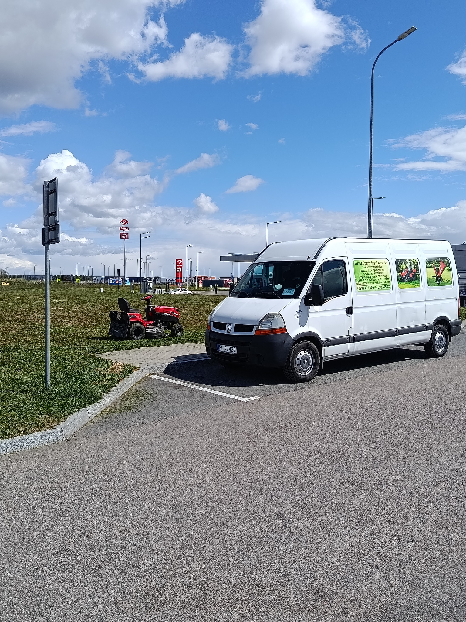 Biały bus z reklamą firmy ogrodniczej i kosiarka samojezdna na trawniku. Usługi: koszenie, przycinanie, wycinanie krzewów, czyszczenie kostki, pranie aut.