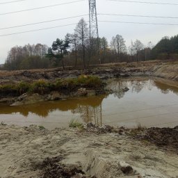 D.DKOP - Wykopany staw z wodą, otoczony ziemią i roślinnością, w tle las i słup wysokiego napięcia. Prace ziemne w terenie podmokłym, widoczne ślady maszyn.
