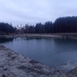 D.DKOP - Szeroki kadr na nowo wykopany staw z brzegami z piasku, otoczony drzewami i budynkami w tle, wykonany w pochmurny dzień. Widoczne ślady prac ziemnych.