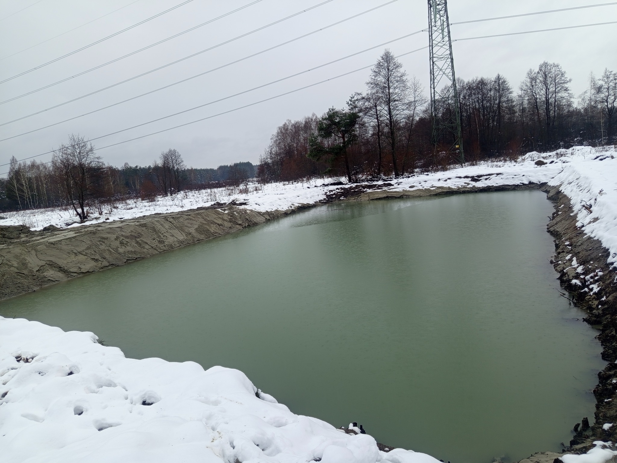 Zimowy krajobraz z wykopanym stawem o mulistej wodzie, otoczonym śniegiem i brzegiem z piasku. W tle las i słup wysokiego napięcia.