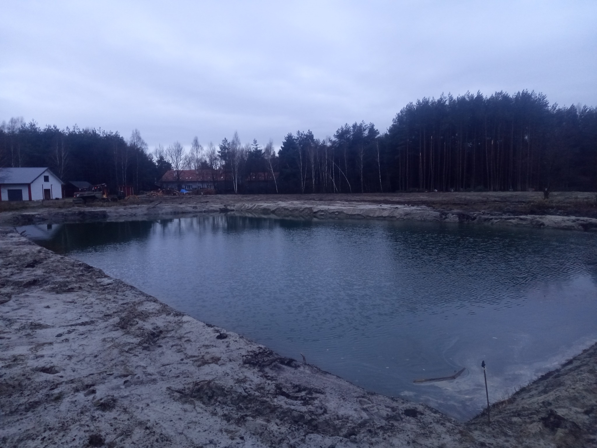 Szeroki kadr na nowo wykopany staw z brzegami z piasku, otoczony drzewami i budynkami w tle, wykonany w pochmurny dzień. Widoczne ślady prac ziemnych.