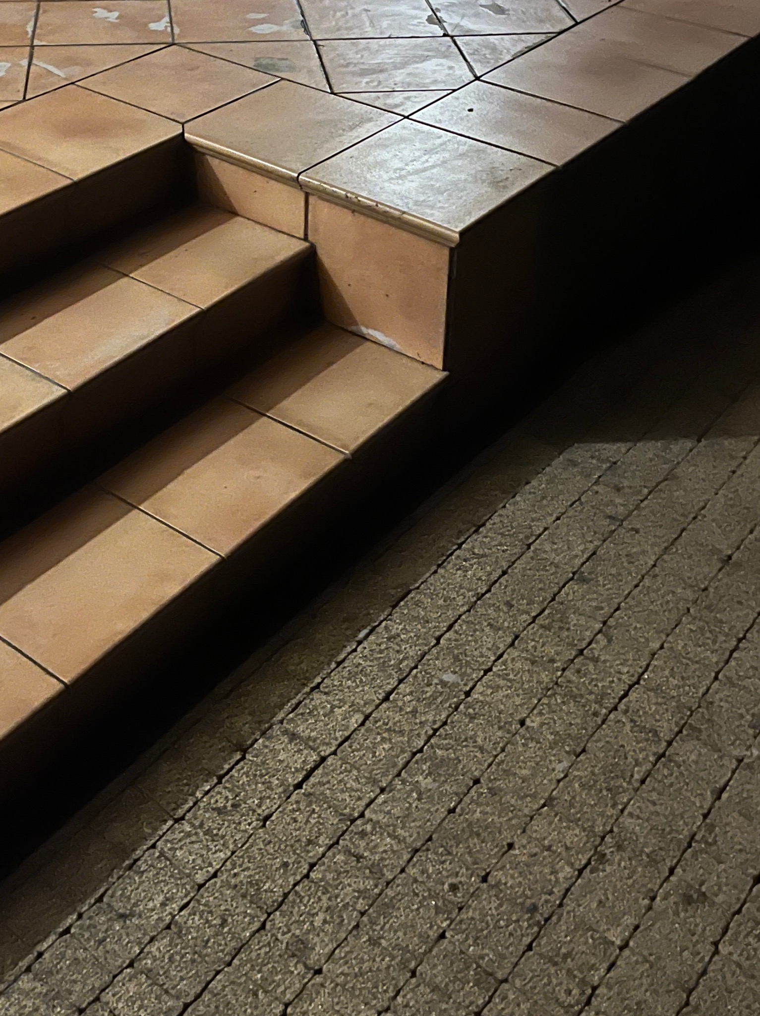 Schody z terakoty przechodzące w kostkę brukową, oświetlone z góry, tworzące kontrast cieni. Widok z góry, podkreślający fakturę materiałów.