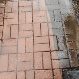 Michał Nowak - Częściowo umyta, mokra kostka brukowa w odcieniach czerwieni i brązu. Widoczna różnica w czystości między umytą a brudną powierzchnią. Woda zbiera się w zagłębieniach.